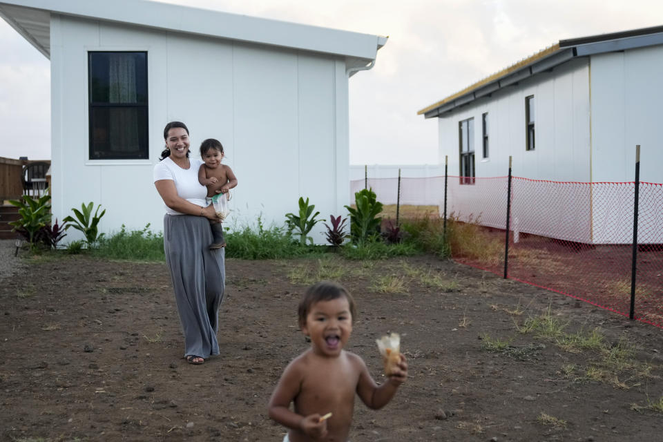 Josephine Fraser, 22, who was born and raised in Lahaina, holds her son Zyon Dias, 18 months, as she watches Ireh Dias, 3, front, run around outside their small home at Ke Ao Maluhia at the Maui Lani housing development, spearheaded by the Council for Native Hawaiian Advancement, Wednesday, July 10, 2024, in Kahului, Hawaii. The family has moved multiple times over the past 11 months after being displaced by the 2023 wildfire and are the first to move into the modular home community being built for those affected. (AP Photo/Lindsey Wasson)