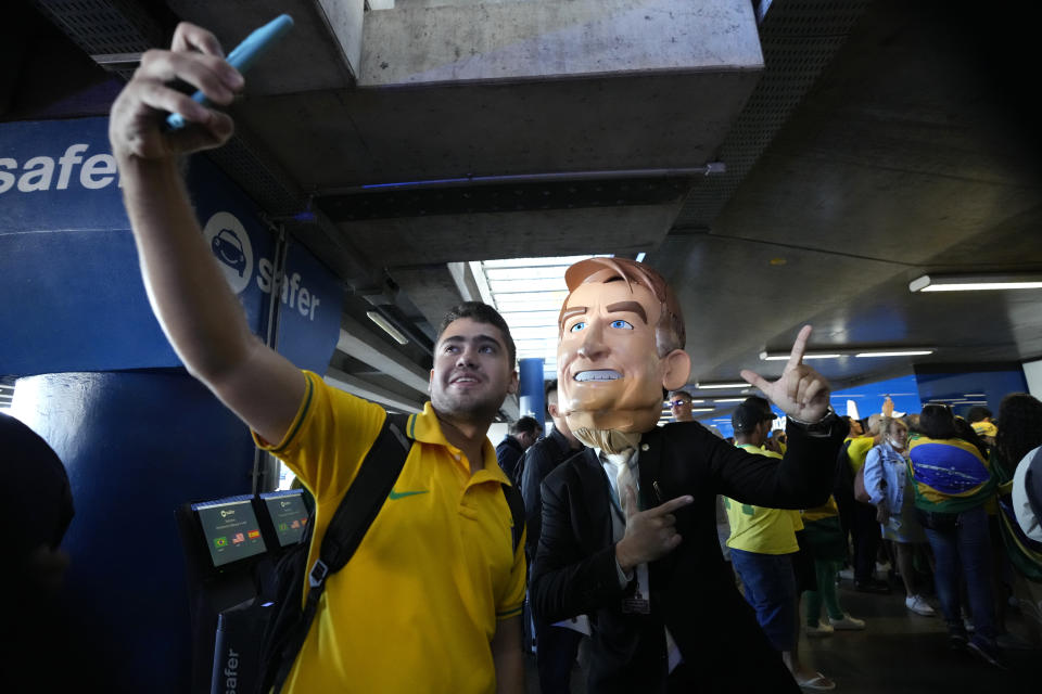 A supporter takes a selfie with a man wearing a mask in the likeness of Brazil's Former President Jair Bolsonaro at the Brasilia International Airport, in Brasilia, Brazil, Thursday, March 30, 2023. Bolsonaro arrived back in Brazil on Thursday after a three-month stay in Florida, seeking a new role on the political scene. (AP Photo/Eraldo Peres)