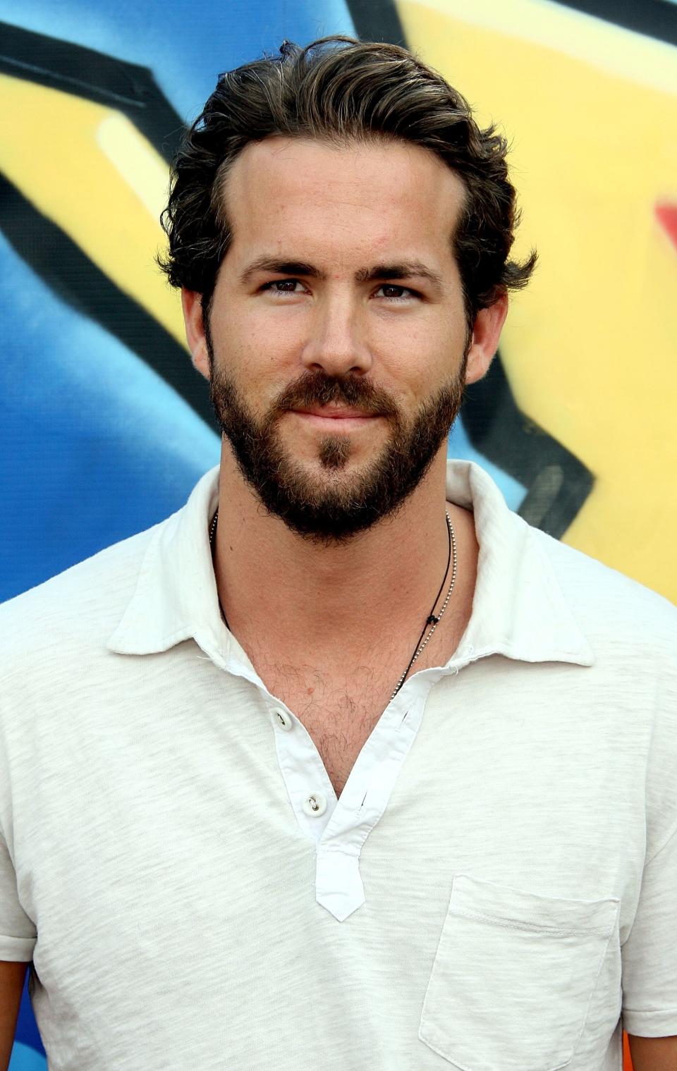 Ryan Reynolds smiling, wearing a casual white shirt with a slightly open collar, standing in front of a colorful background
