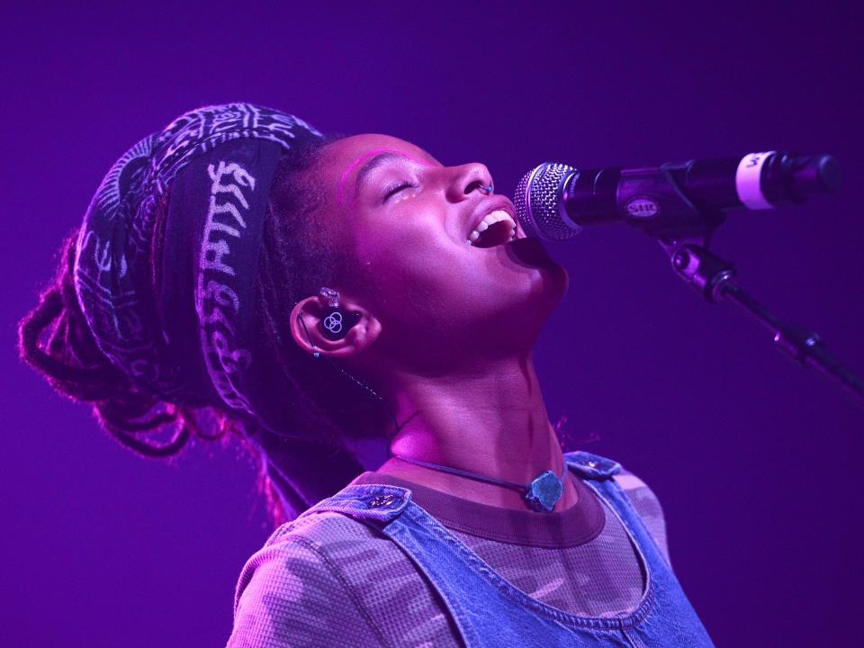 Profile of WILLOW singing into a mic, facing towards the sky. Her hair is loosely wrapped up in a patterned cloth. She's wearing a henley, overalls, and a turquoise necklace.