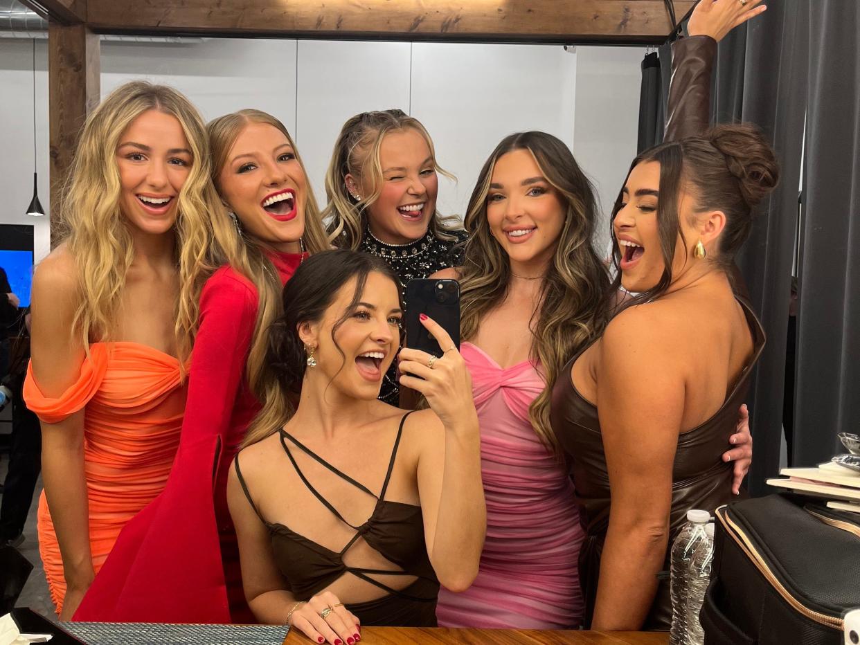 chloe, paige, jojo, kendall, kalani, and brooke from dance moms posing together as young aults. they're all young women wearing dresses, posing together for a selfie in a production studio