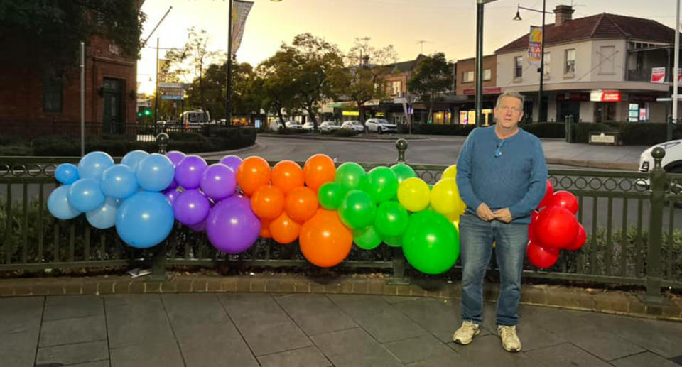 A man can be seen in jeans and a blue jumper standing in front of the balloons. 