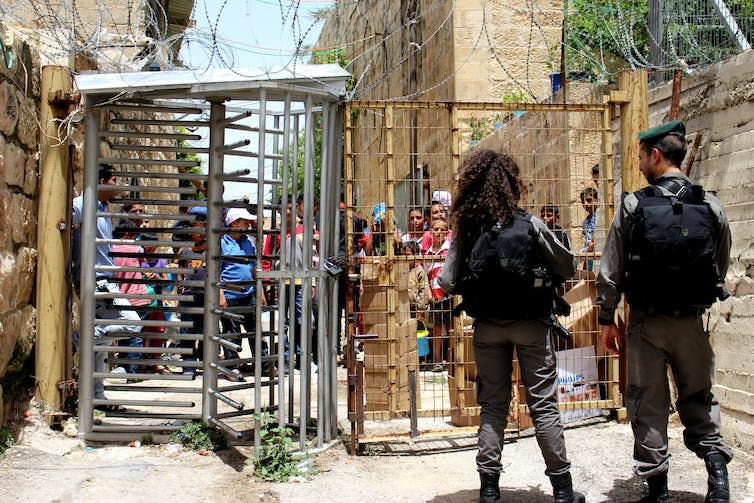 Palestinian children wait at a security checkpoint in Hebron, guarded by Israeli soldiers.