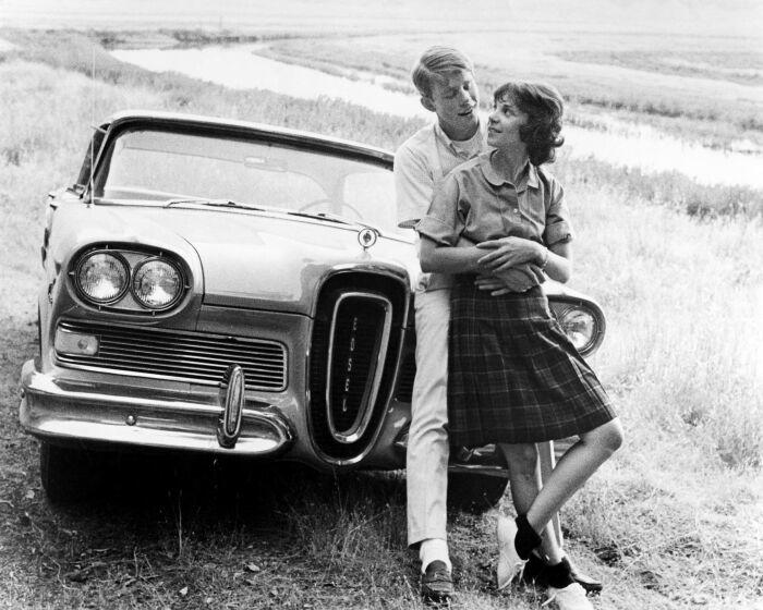 A black and white photo of a man holding a woman in front of a vintage car