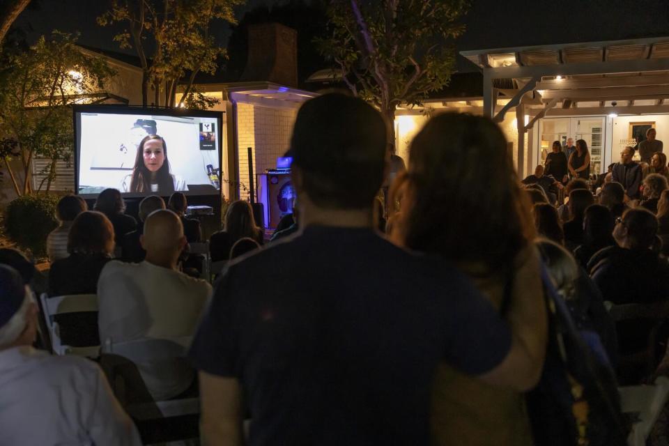 A large screen shows a woman while a crowd watches outdoors.