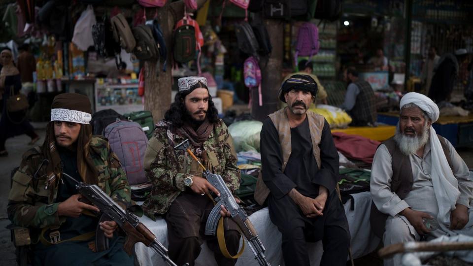 Taliban-Kämpfer sitzen neben Straßenverkäufern auf einem lokalen Markt und halten dabei Waffen in den Händen.