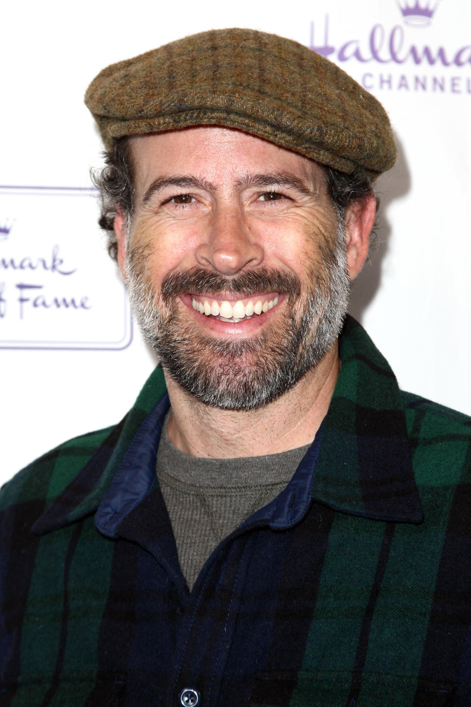 Jason Lee smiles while wearing a plaid shirt, grey undershirt, and a flat cap at a Hallmark Channel event