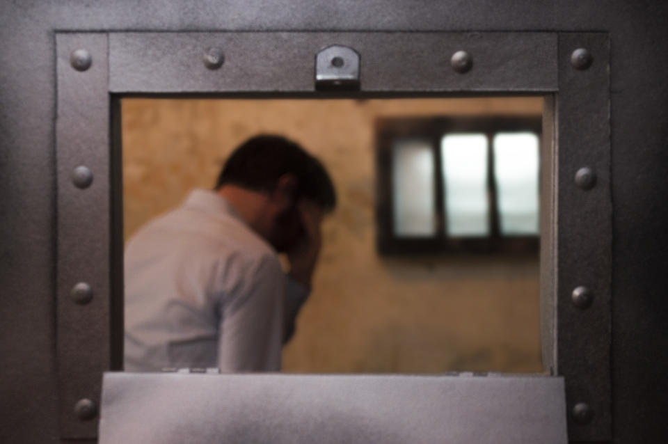 A man stands in a prison cell by a barred window, his head in his hand.