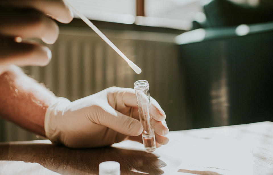 A hand wearing a latex glove holds a small clear tube and dips a cotton swab into the liquid. Space for copy.