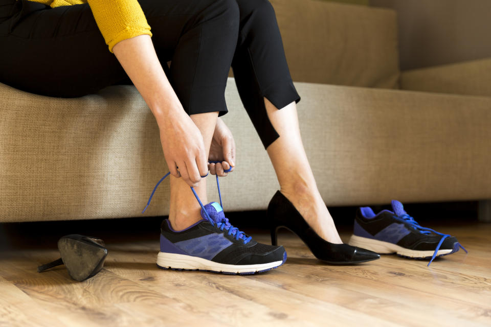 A woman lacing up a sneaker on her right foot while wearing a high heel on her left foot.