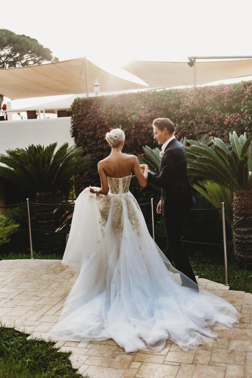 A groom helps a bride walk down a hill in a wedding dress.