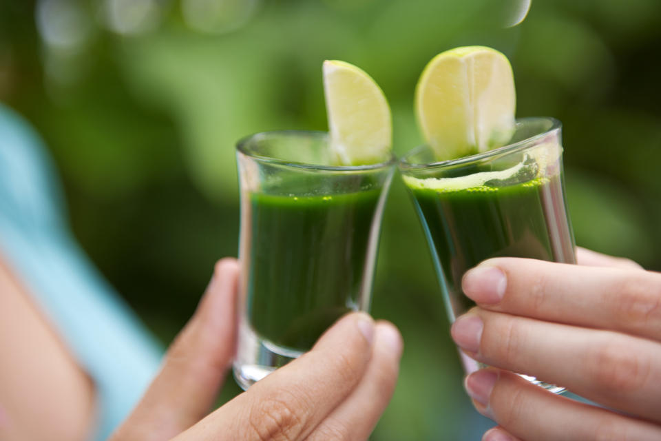 Photograph of two female hands holding two shots of wheatgrass with lime and cheering 