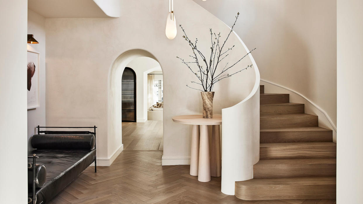  Minimalist hallway with white textured walls, sweeping curved wooden staircase, black leather daybed and off-white travertine round table 