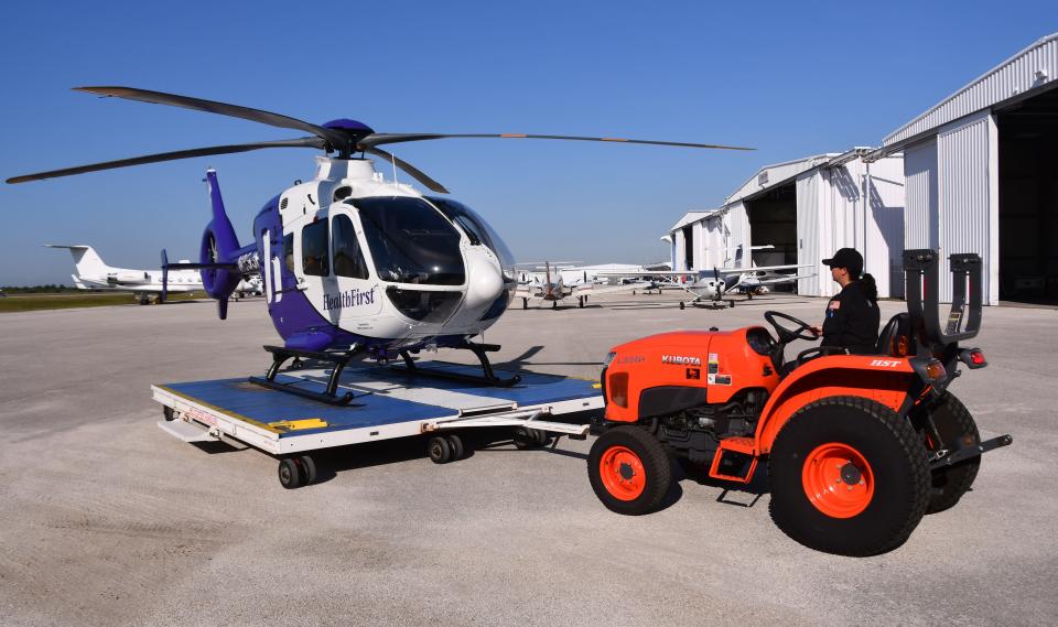 An orange Kubota tractor pushes and pulls the helicopter in and out of the hangar using a wheeled platform.