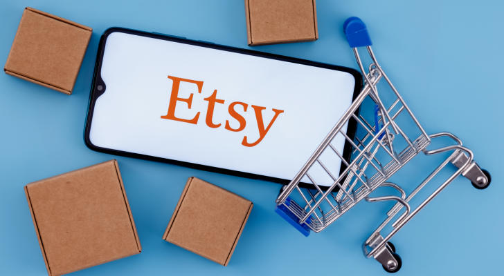Etsy logo on a phone screen on a blue background. Phone is in a little cart and there are packages around them. ETSY stock.
