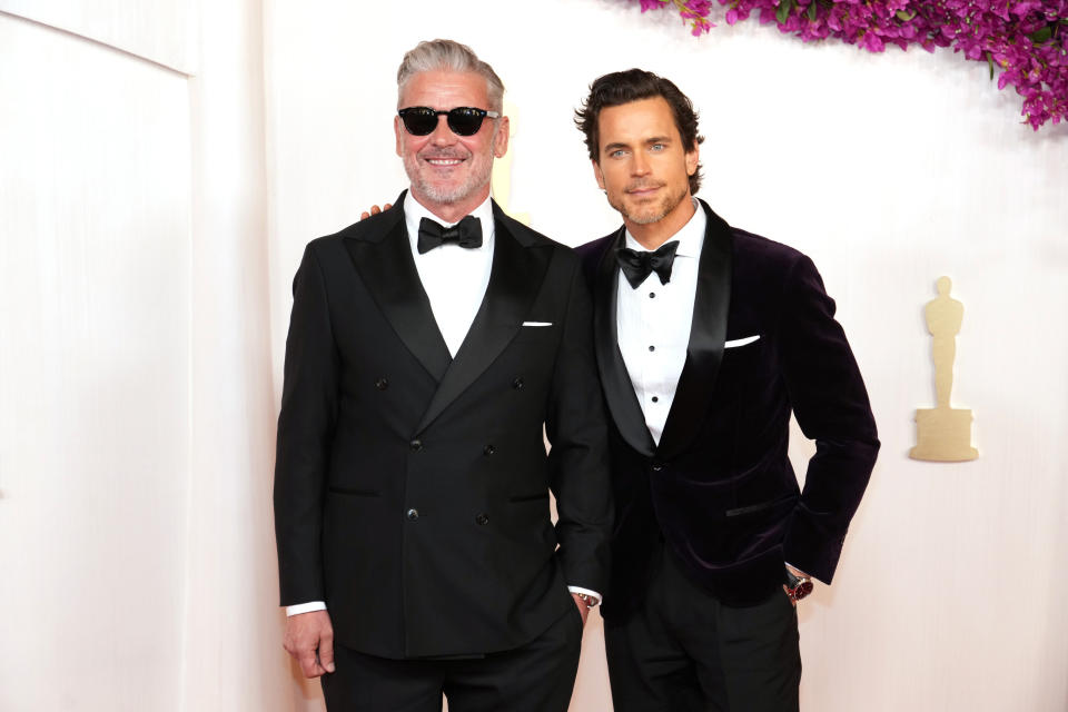 Simon Halls and Matt Bomer pose together in formal attire at a red carpet event. Simon wears a black tuxedo with sunglasses; Matt wears a velvet tuxedo jacket