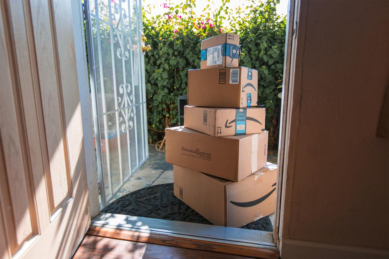 Five Amazon boxes on doorstep of home with open front door, flowering hedges in the background