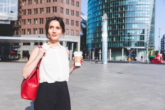 A woman in a sleeveless blouse and dark pants stands outdoors with a red handbag, holding a coffee cup, with modern buildings visible in the background