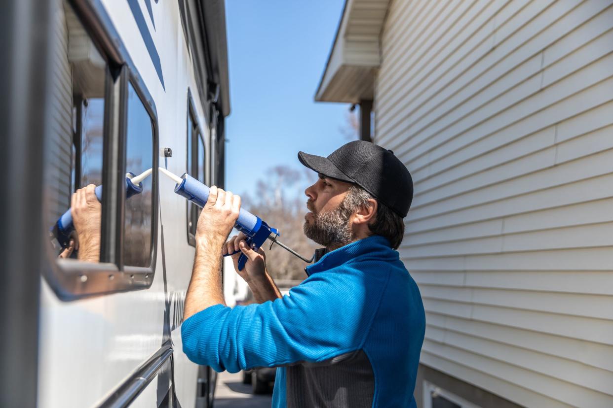 man is doing the maintenance of a camper trailer. He is applying a sealant around the windows and other parts of the trailer. 