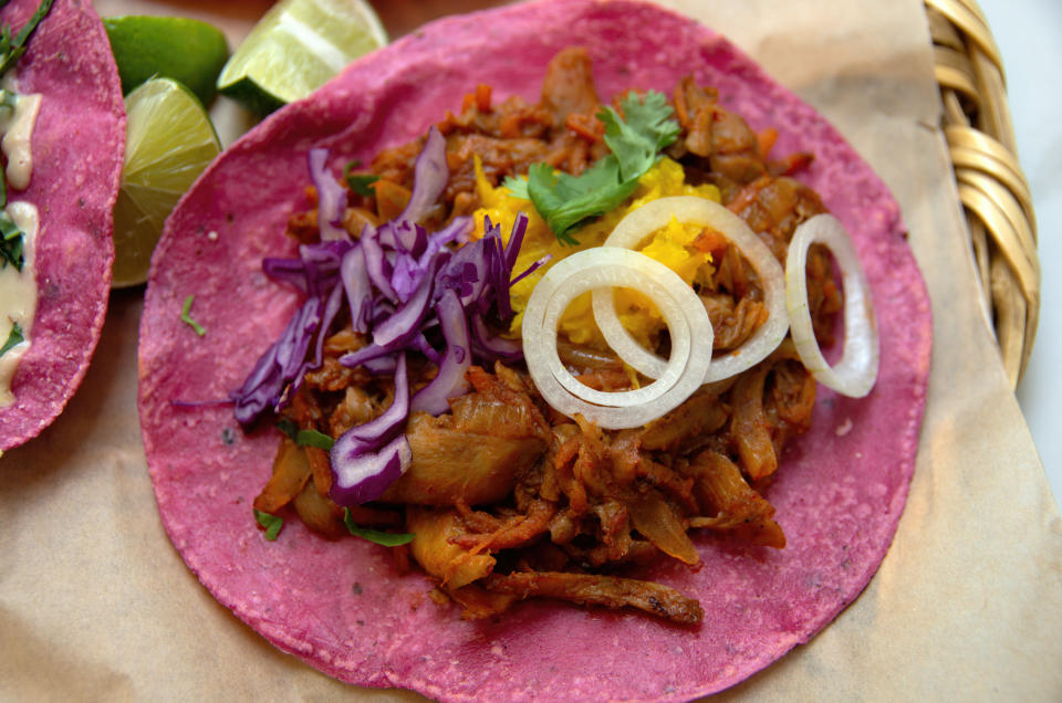 Taco with onions, cilantro, and lime on a pink tortilla; additional taco and lime slice in background