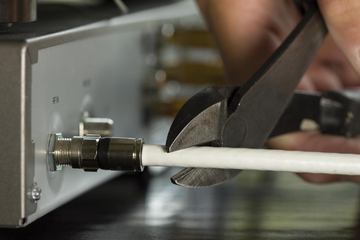 Closeup of hand holding pliers cutting a white cable TV cable that is attached to the back of a cable box with the rest of the cable box blurred in the background