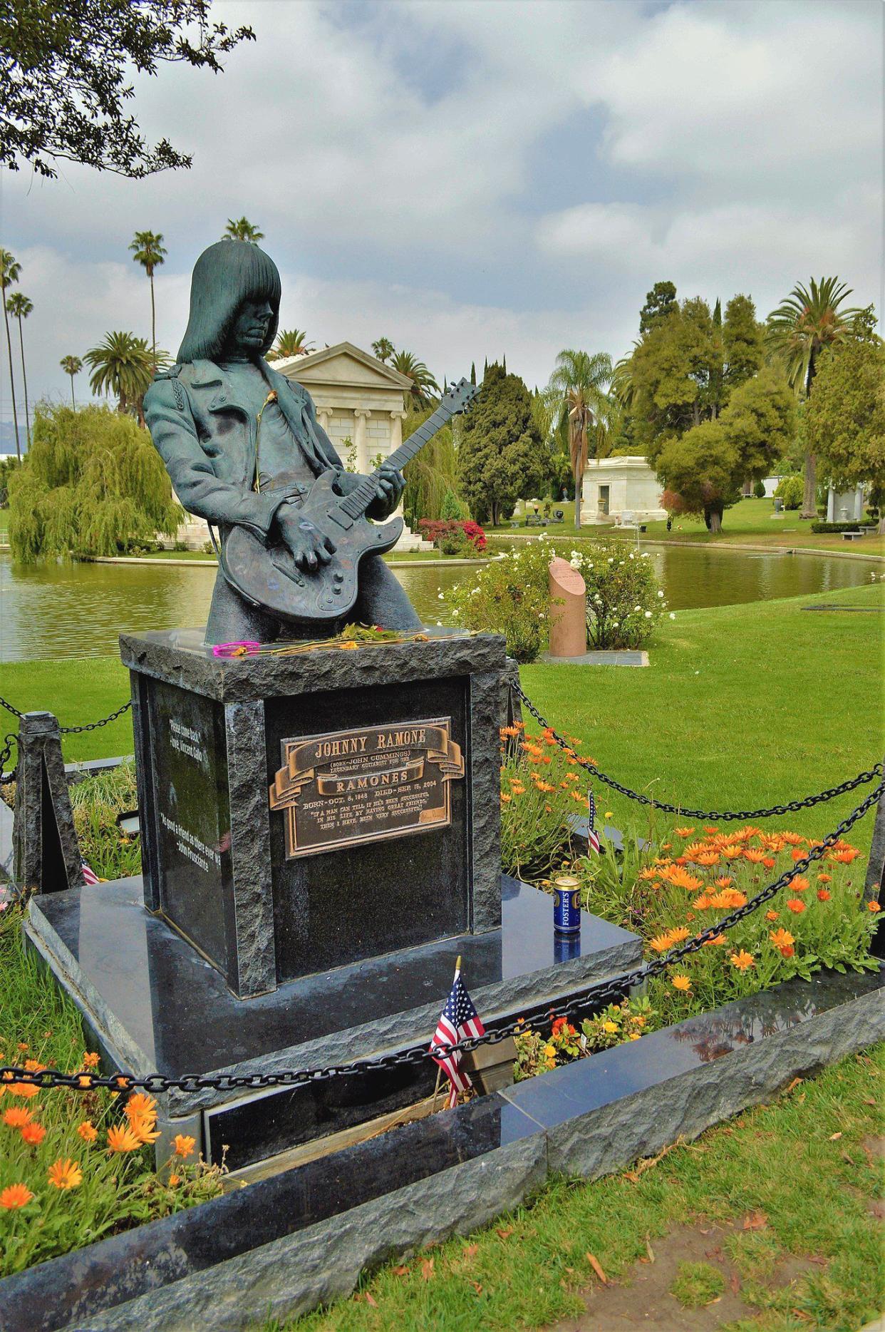 Johnny Ramone's statue and grave in the Hollywood Forever Cemetery, Los Angeles surrounded by orange flowers in bloom and black stone with a lake and several other tombs in the background