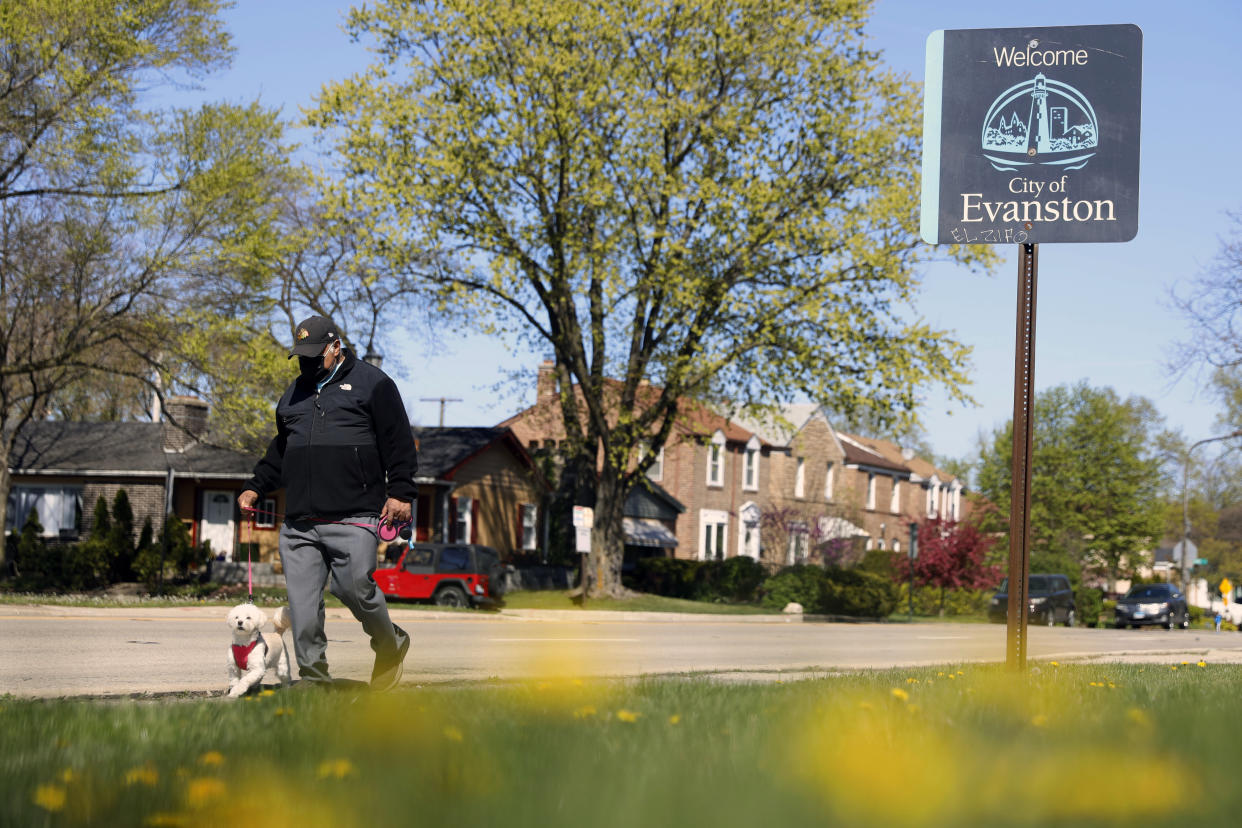 An older man in a baseball cap walks a small white dog past a street sign reading: Welcome City of Evanston,