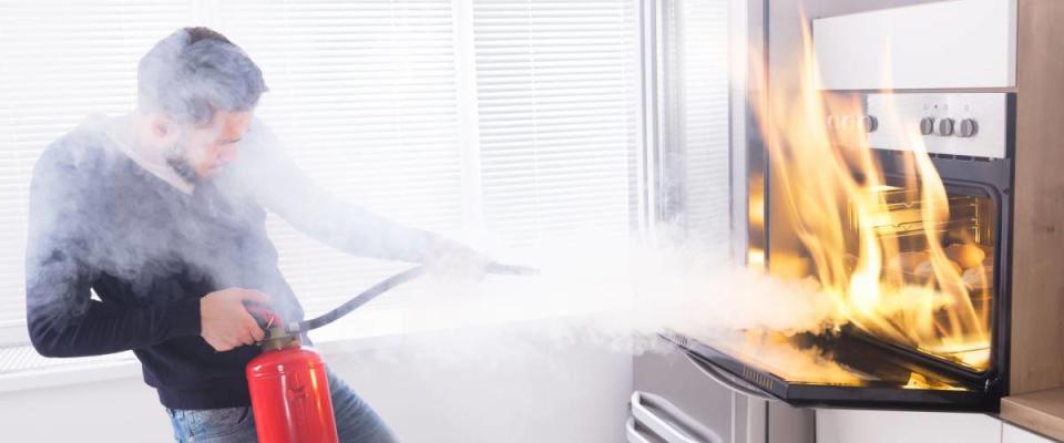 Young Man Using Red Fire Extinguisher To Stop Fire Coming From Oven In Kitchen