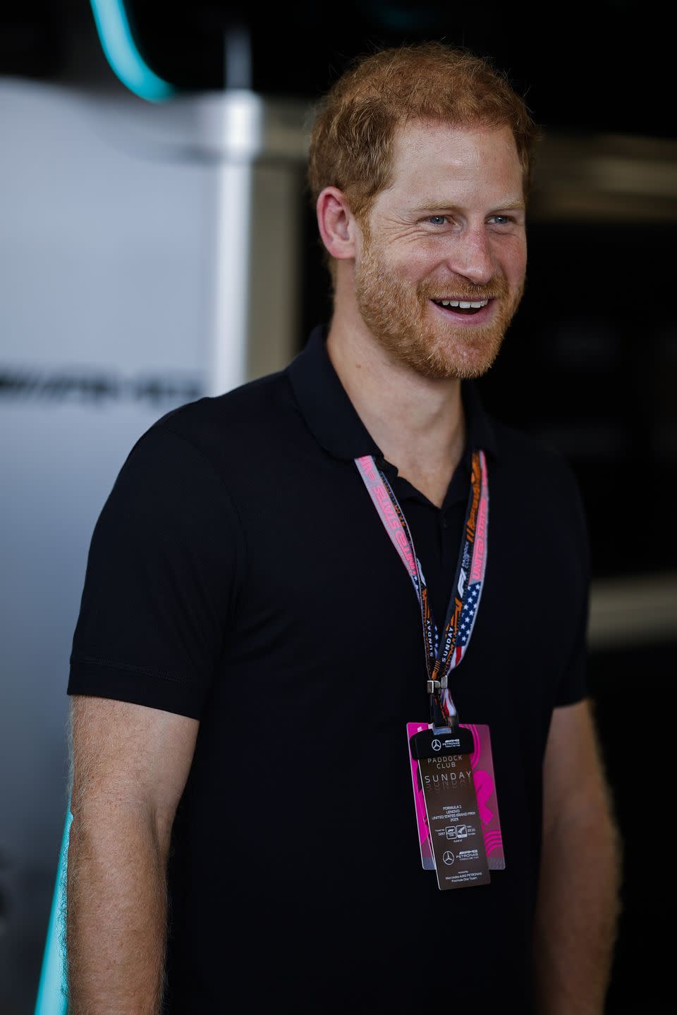 prince harry smiles and looks right, he wears a black polo shirt and two lanyards around his neck with cards on the bottom
