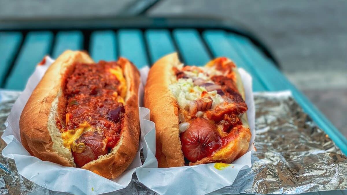 Chili Man's Choice and Southern All-The-Way Hot Dogs on individual white wrapping papers on aluminum foil, The Chill Man, Charlotte, on a blue outside seat blurred in the background