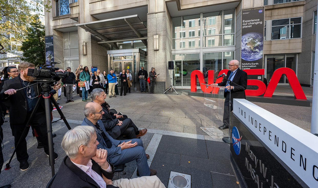  A man in a blue suit speak in an outdoor courtyard with "nasa" written in red in the background. 