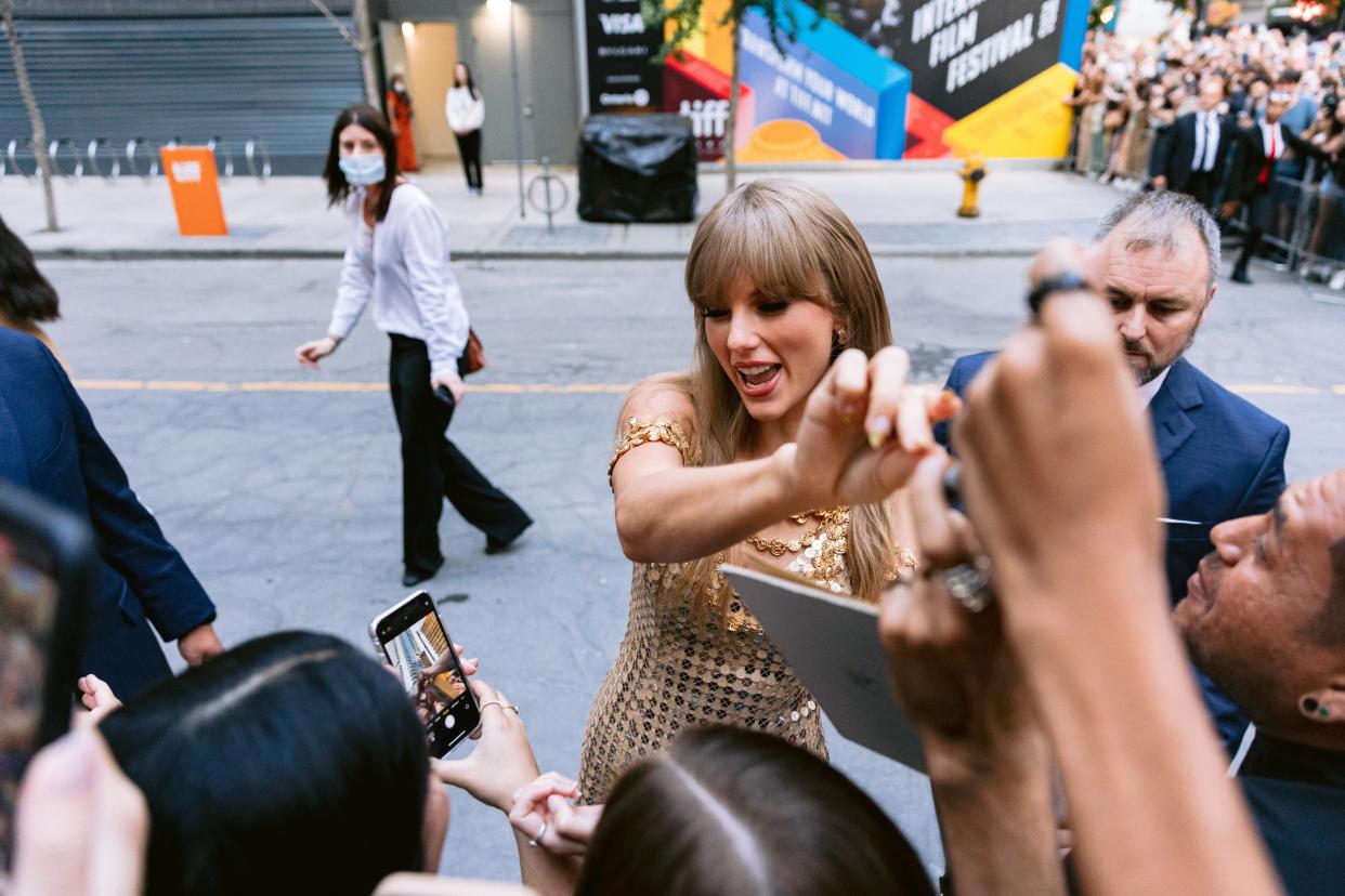 Taylor Swift greets fans on a sidewalk while wearing a golden dress.