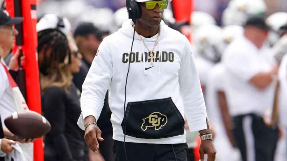 deion sanders wearing sunglasses and a headset as he walks the sideline during a football game