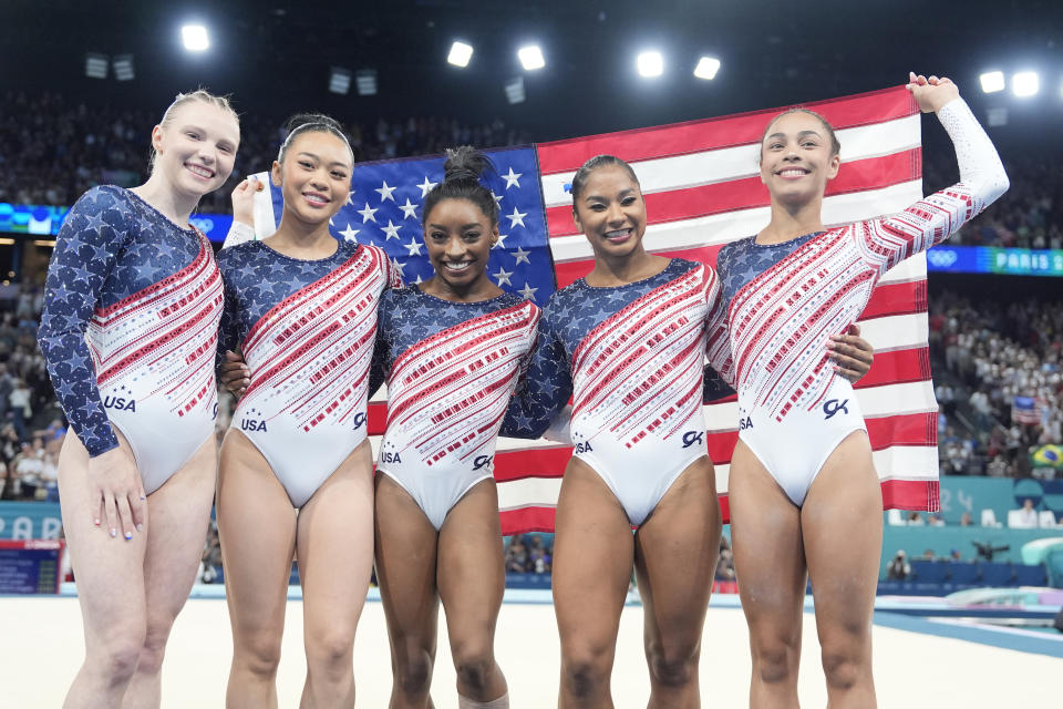 Team USA, from left to right – Jade Carey, Suni Lee, Simone Biles, Jordan Chiles and Hezly Rivera – celebrates gold while holding the U.S. flag. 