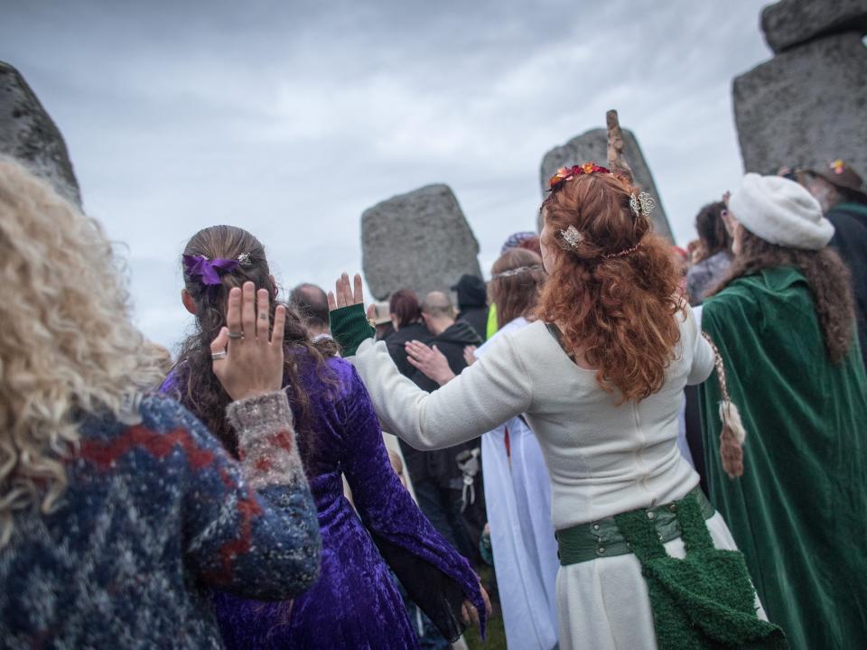 People, some in traditional garb, hold their hands up towards the sky, with Stonehenege in the background.