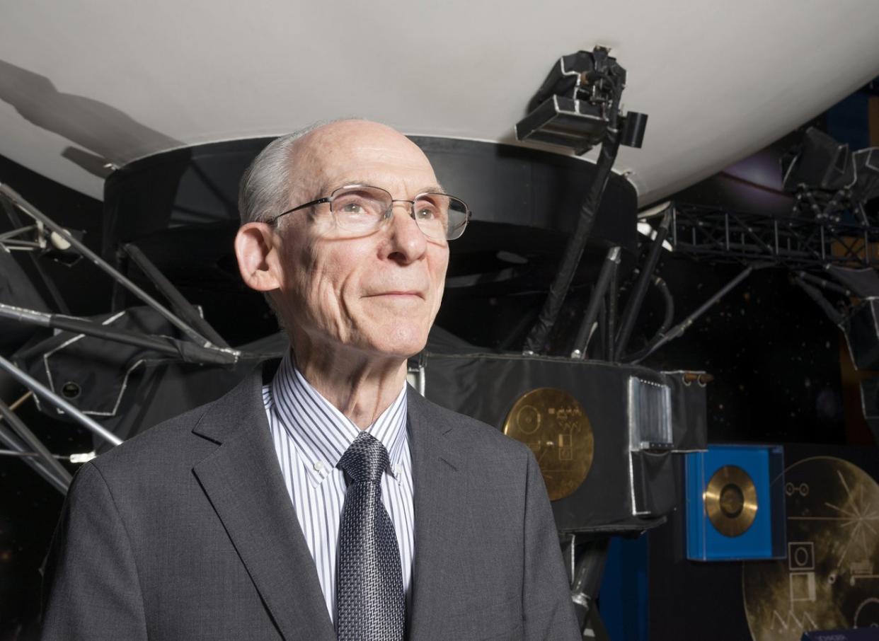  A smiling older man in a suit stands in front of a model of a large spacecraft. . 