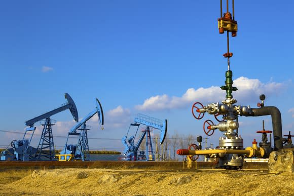 A close-up of a natural gas wellhead with three oil pumps in the background.