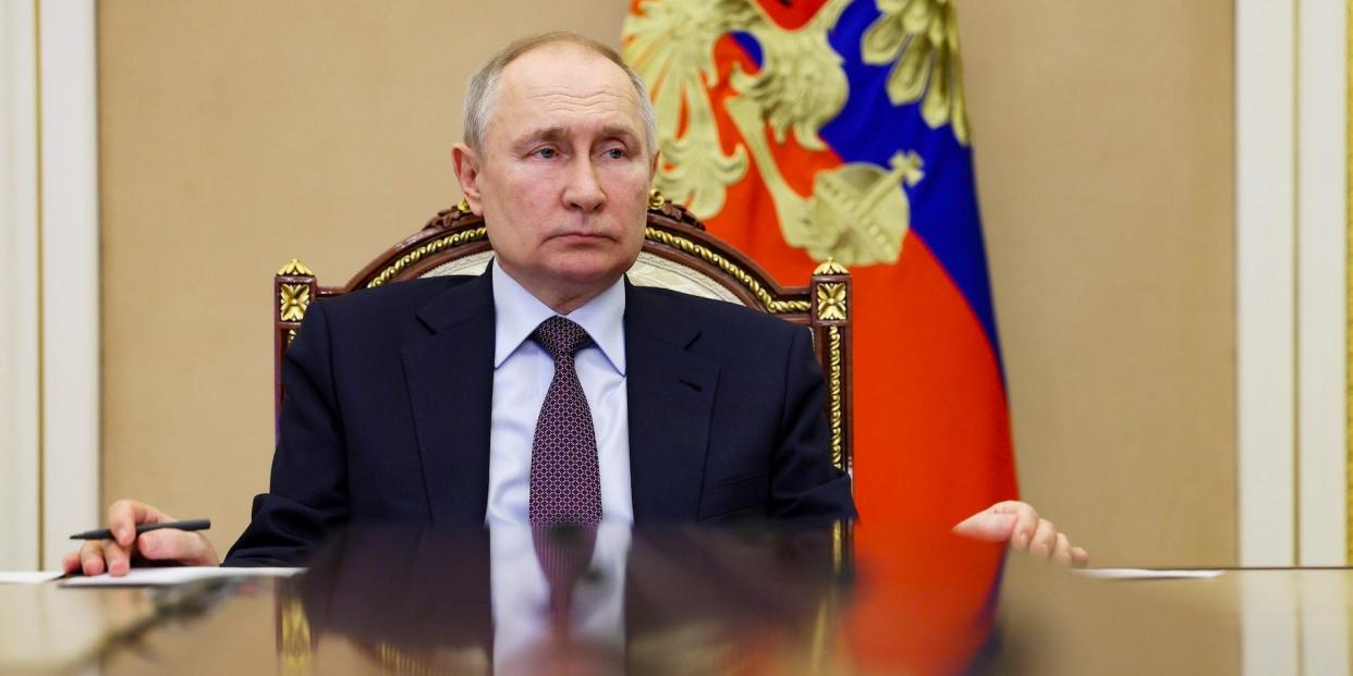 Russian President Vladimir Putin sitting in a chair in front of a Russian flag.