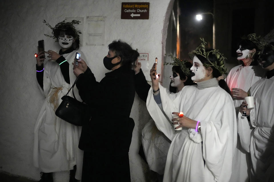 A woman and participants with faces painted wear white sheets and hold torches on long poles, take photos of the Torch Parade (Lampadiforia) on the Aegean Sea island of Naxos, Greece, late Saturday, Feb. 25, 2023. The first proper celebration of the Carnival after four years of COVID restrictions, has attracted throngs of revellers, Greek and foreign, with the young especially showing up in large numbers. (AP Photo/Thanassis Stavrakis)