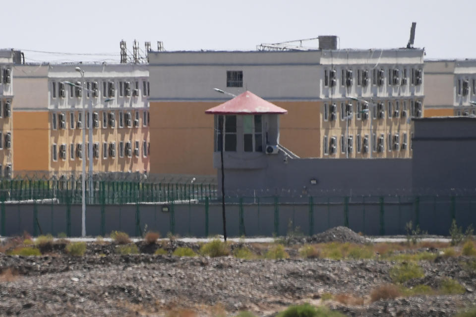 Buildings with bars on windows behind a razor-wire-topped fence and a high wall with guard tower.
