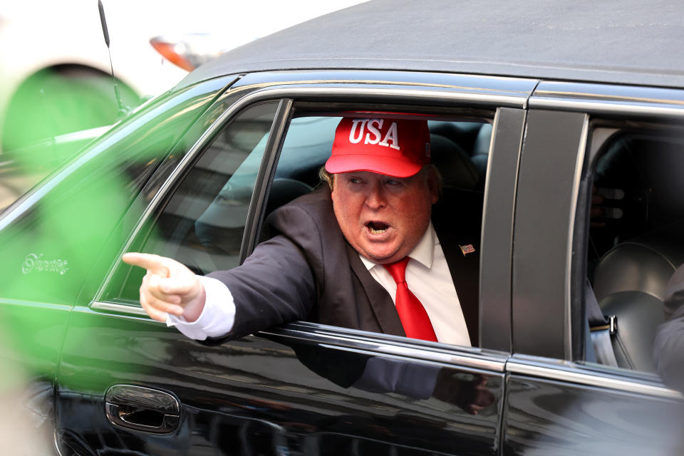 A person dressed as Trump leans out of a car, speaking and pointing his finger, as he rides past Trump Tower.