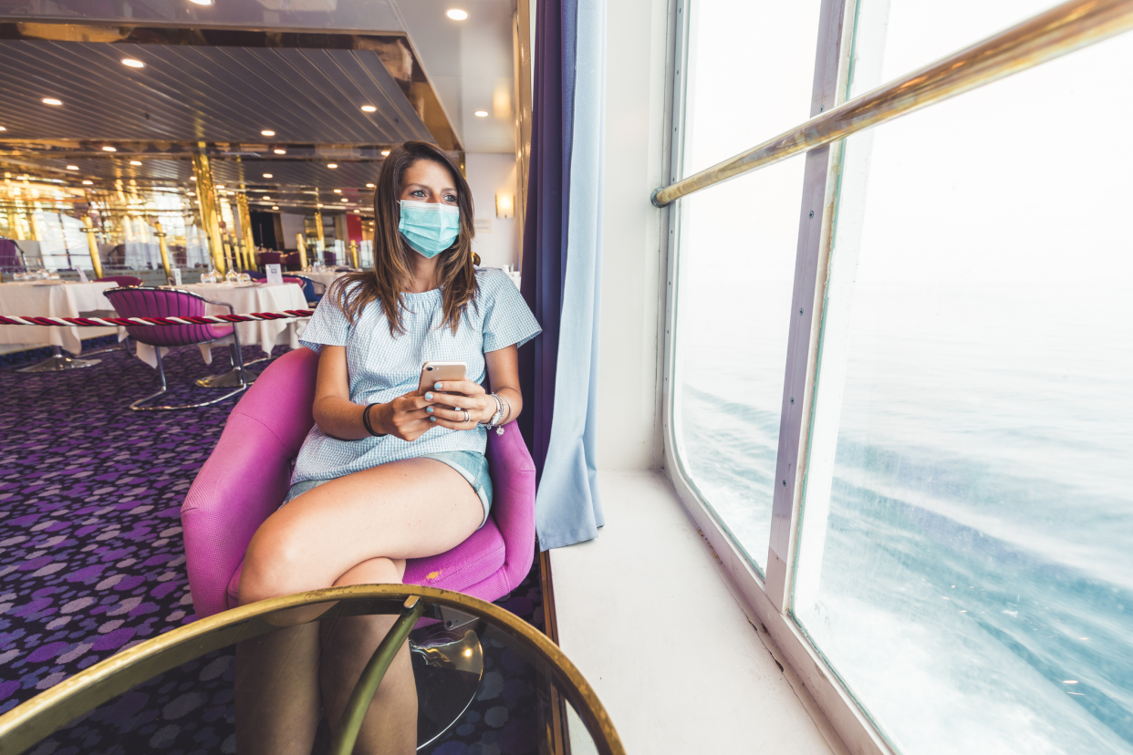 Woman wearing a face mask, siting inside looking out of a window of the ocean, on a cruise in the Mediterranean Sea, dining area in the background