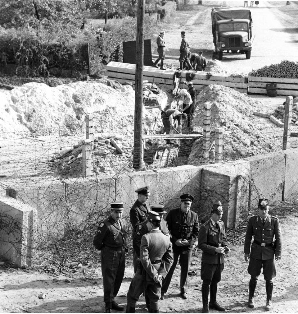 In the foreground, French Military Police, left, West Berlin alert police, right, and West Berlin custom officials are on guard in front of the concrete wall on the western side of the Berlin Wall in the French sector near Schoenholz, Germany, on Sept. 27, 1961. In the background behind the concrete wall and barbed wire fence are East Berliners working on cable. Beyond them East German soldiers guard the border.