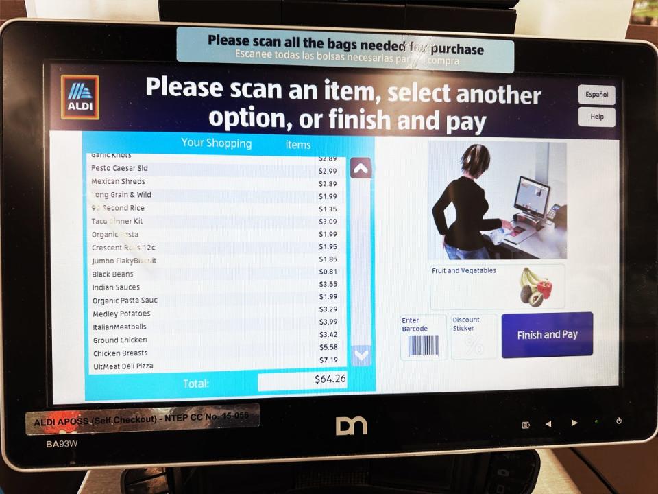 Screen at Aldi self-checkout station. The screen shows an illustration of a woman at a self-checkout counter and a list of several scanned items and a total bill of $64.26