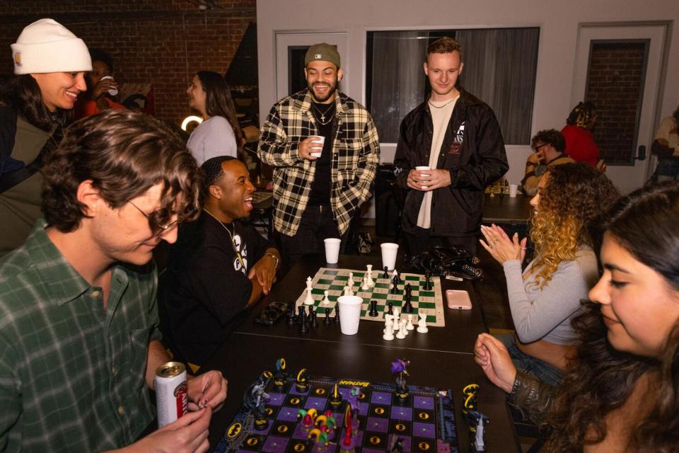 A group of people, some sitting and some standing, playing chess and socializing.