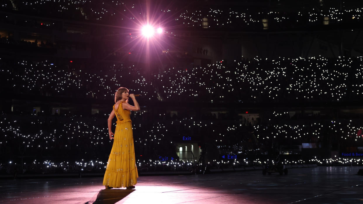  Taylor Swift on stage in a yellow dress for her Eras Tour performance with a purple spotlight on her. 