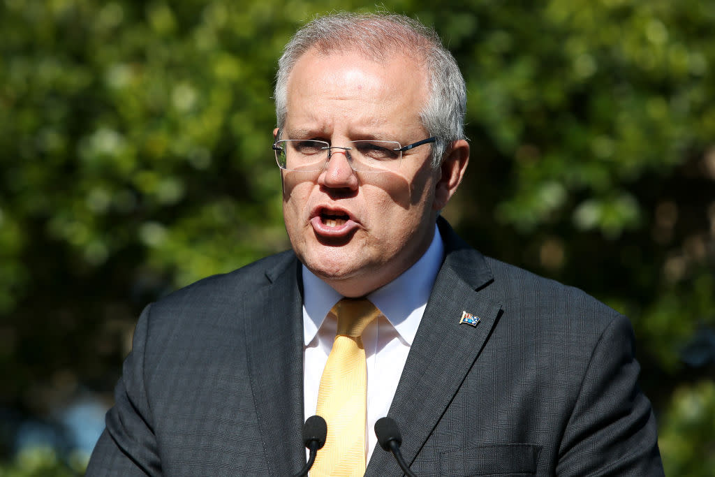 A close up of Scott Morrison speaking. He wears a grey suit jacked with a yellow tie. He is wearing glasses, and the photo is taken mid-speech. 