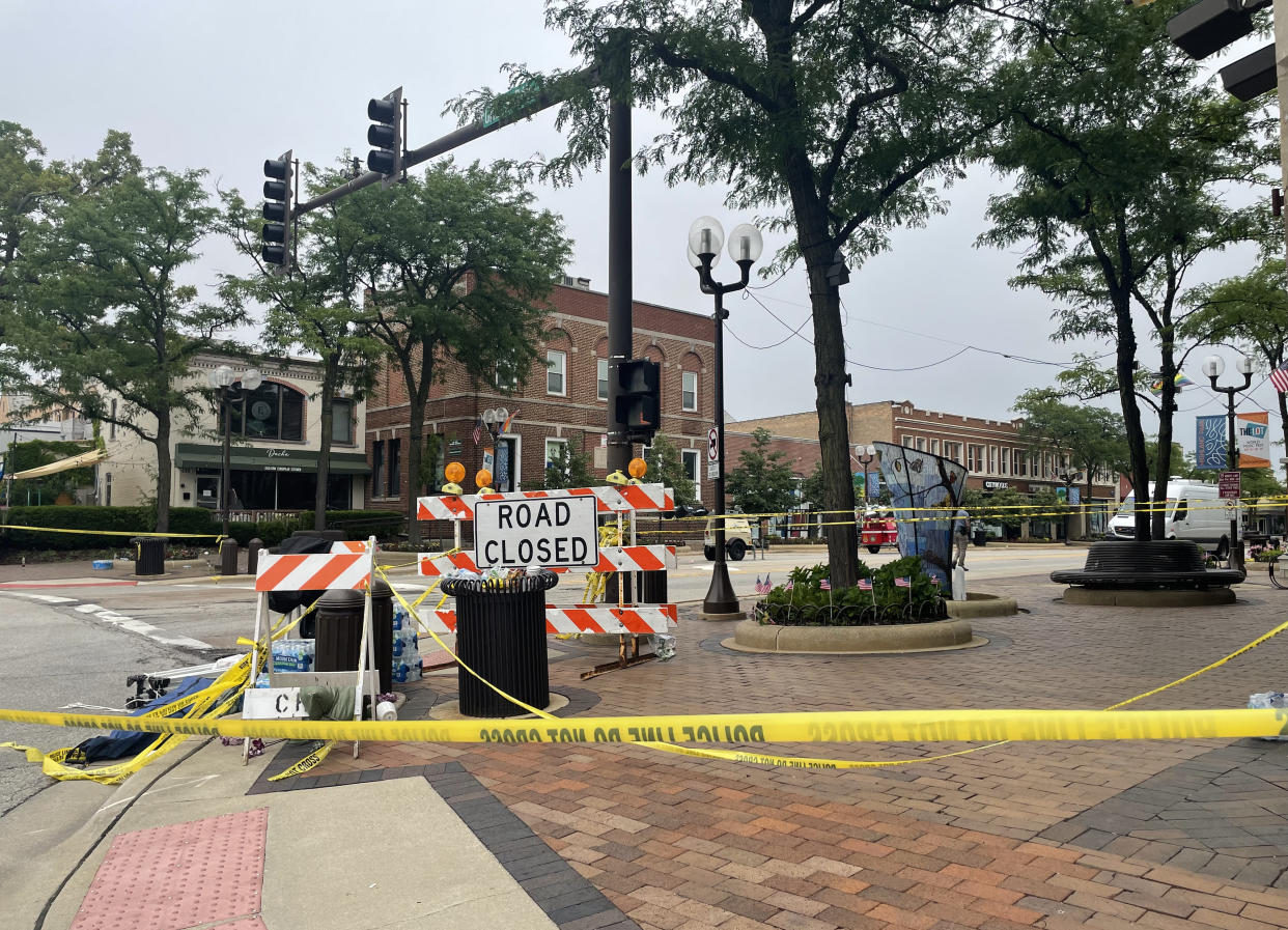 Yellow police tape cordons off a brick-sidewalk-lined intersection with sign reading Road Closed.