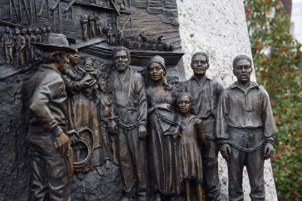 Six figures of enslaved people are rendered in brass. Three are men; two are women; one is a child. The men wear shirts and overalls; the women dresses and headscarves; the child a dress. Three men and a women are visibly chained to each other. In the foreground, a bearded white man wearing a fedora and holding a gun and a whip looks at the enslaved individuals.