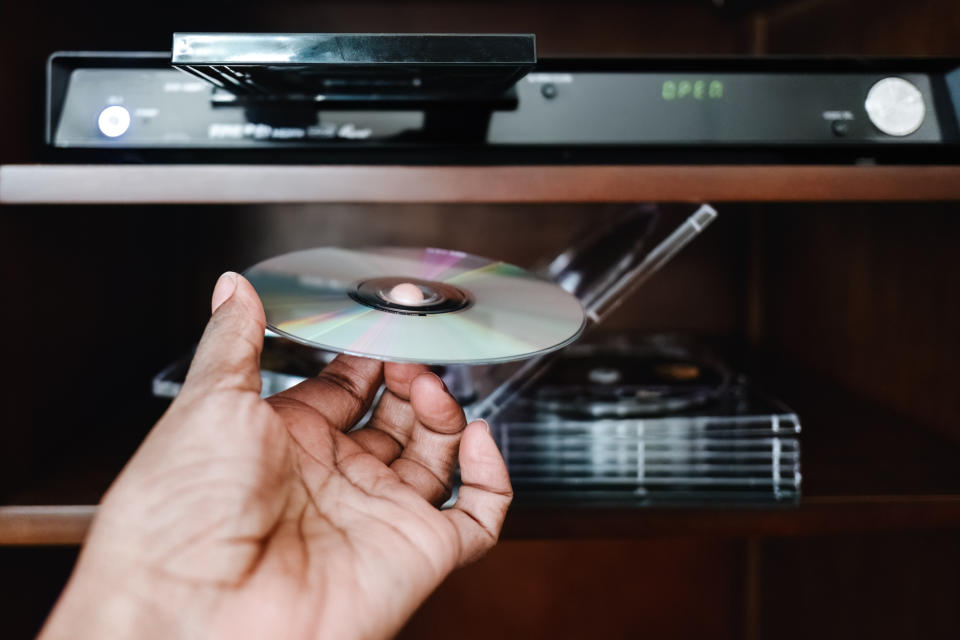 A hand inserting a DVD into a DVD player shelf, with several other DVDs visible. The DVD player is turned on